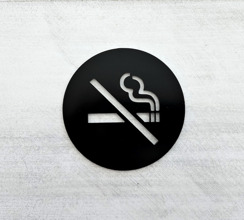 May include: Black and white no smoking sign with a cigarette and smoke crossed out. The sign is round and has a white background.