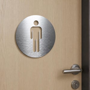 May include: A circular brushed metal sign featuring a die-cut silhouette of a person. The sign has a smooth, metallic finish with horizontal graining and is mounted on a light-colored wooden door with a silver-toned metal door handle and lock.