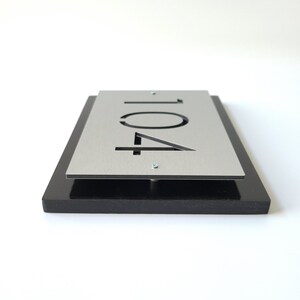May include: A silver metal house number plaque with the number "104" cut out. The plaque is mounted on a black wooden base.