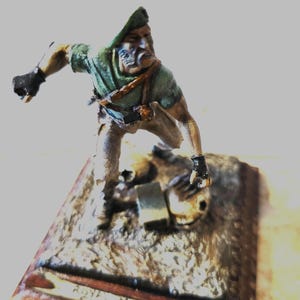 Può includere: Figurina in miniatura di un Royal Marine in uniforme verde e basco, in posa d'azione. La figurina è montata su una base rettangolare con la scritta "ROYAL MARINE 1982".
