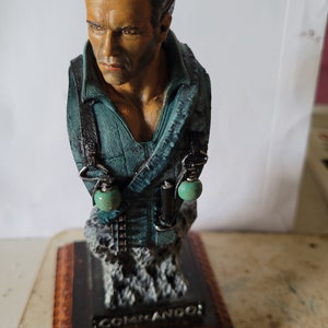 May include: A detailed bust of a man with dark hair and a stern expression, wearing a teal jacket with black straps. The bust holds two green grenades. The base reads "COMMANDO" and "ARNOLD SCHWARZENEGGER".