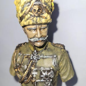 Puede incluir: Busto detallado de una figura militar, con un sombrero amarillo y marrón con un emblema de calavera. La figura lleva un uniforme verde con detalles dorados y plateados, incluyendo una medalla en forma de cruz. El busto está sobre un fondo neutro.