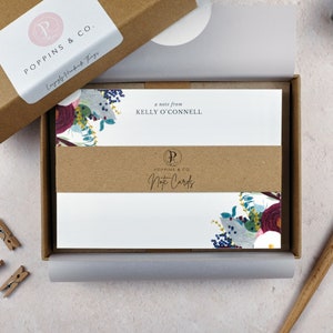 May include: A box of note cards featuring a floral design in shades of blue, purple, and white. The box is brown and has a label that reads "Poppins & Co. Note Cards".