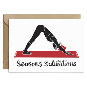 May include: A greeting card featuring a black silhouette of a person in a downward-facing dog yoga pose, wearing a Santa hat, on a red yoga mat. The words "Seasons Salutations" are written in black script below the image. The card is inside a brown envelope.