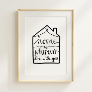 May include: A framed art print featuring a black line drawing of a house shape with a heart above it. The words "home is wherever I'm with you" are written in a cursive font within the house outline. The frame is a light wood color.