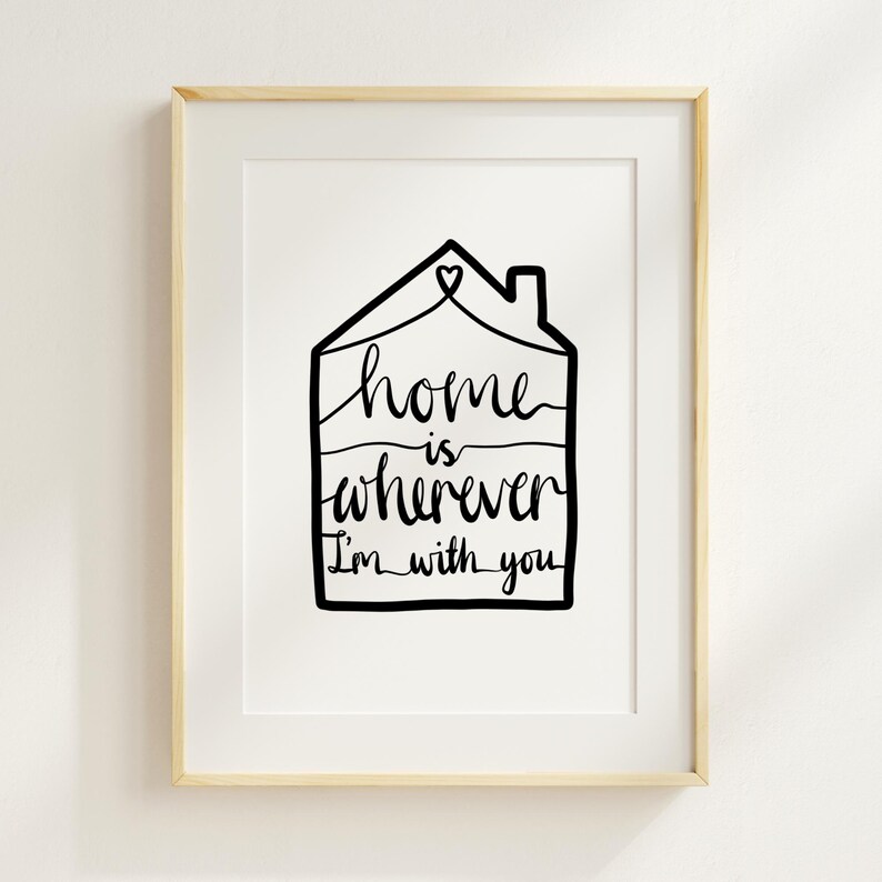 May include: A framed art print featuring a black line drawing of a house shape with a heart above it. The words "home is wherever I'm with you" are written in a cursive font within the house outline. The frame is a light wood color.