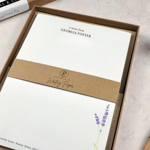 以下が含まれることがあります： 「a letter from GEORGIA FOSTER」の文字とラベンダーのイラストが入った白い便箋のセット。紙は「POPPINS & Co. Writing Paper」と書かれた茶色のバンドが付いた茶色の箱に入っています。