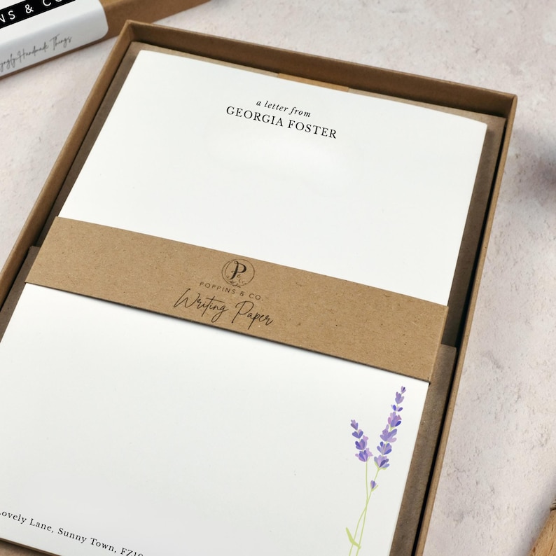 以下が含まれることがあります： 「a letter from GEORGIA FOSTER」の文字とラベンダーのイラストが入った白い便箋のセット。紙は「POPPINS & Co. Writing Paper」と書かれた茶色のバンドが付いた茶色の箱に入っています。