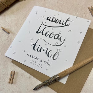K&ouml;nnte beinhalten: Eine wei&szlig;e Verlobungskarte mit schwarzer Kalligraphie, die "about bloody time" liest. Die Karte enth&auml;lt die Namen Harley & Tom, das Wort "ENGAGED" und das Datum 09.03.2024. Kleine Herz-Kritzeleien und eine Feder sind ebenfalls sichtbar.
