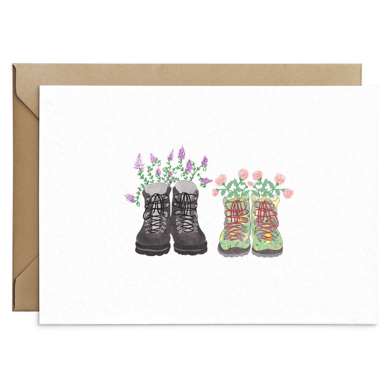 May include: Greeting card featuring watercolor illustrations of hiking boots. One pair is black with purple flowers, the other is green with pink roses. The card is on a white background with a brown envelope.