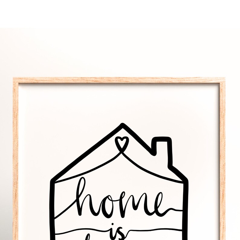 May include: A framed print with the words "home is" in a handwritten font, inside a black house outline with a heart. The frame is a light wood color, and the background is white.