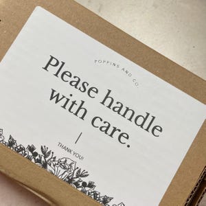 May include: A white label with the text "Please handle with care" in a black, bold font, with the words "POPPINS AND CO." above the text. The label is on a brown cardboard box, with a floral design at the bottom.