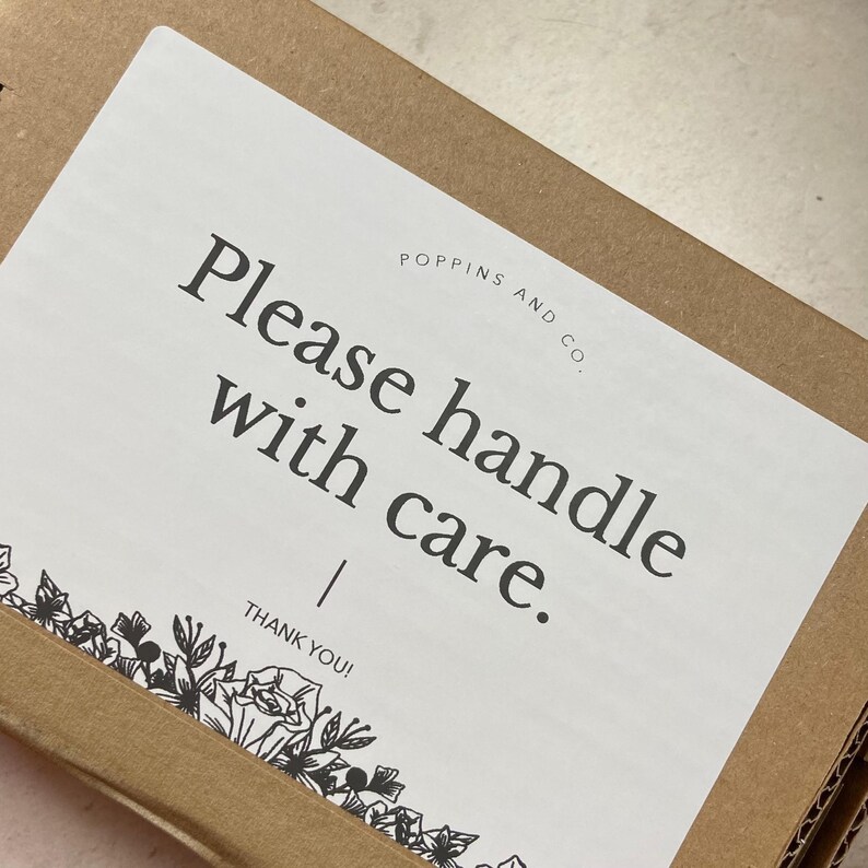 May include: A white label with the text "Please handle with care" in a black, bold font, with the words "POPPINS AND CO." above the text. The label is on a brown cardboard box, with a floral design at the bottom.