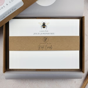 May include: A box of note cards with a brown paper band. The band has a logo that says "Poppins & Co. Note Cards" and a bee illustration. The note cards have a bee illustration and the text "a note from Jolie Juniper Bee".