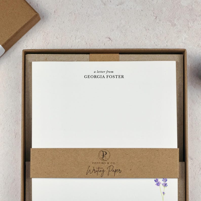 以下が含まれることがあります： 「a letter from GEORGIA FOSTER」と「Writing Paper」の文字が入った白い便箋が入った茶色の箱の文房具セット。セットには「POPPINS & CO.」のロゴが入った茶色のバンドが含まれています。