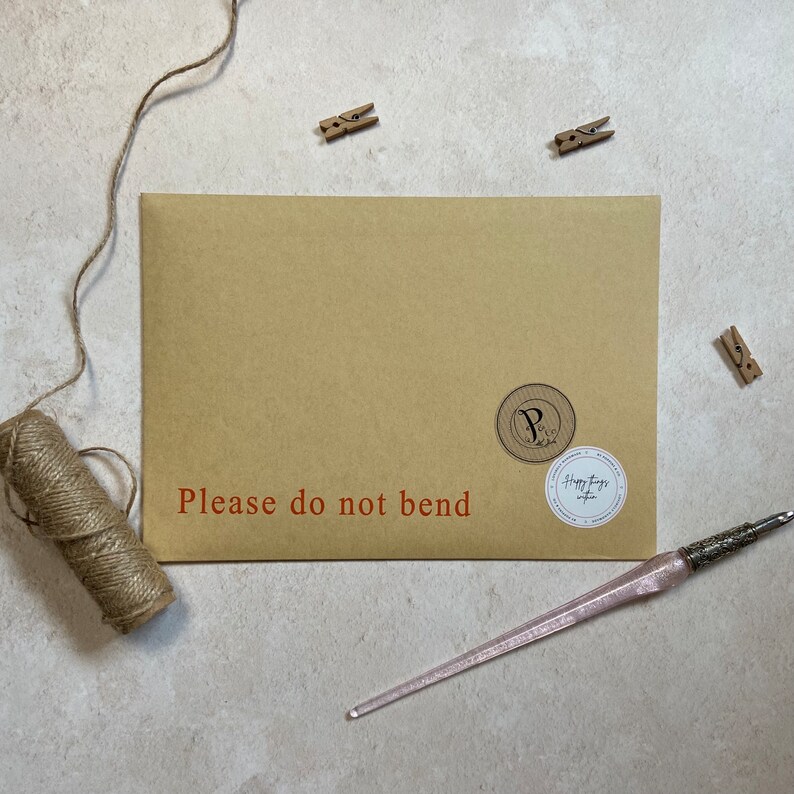 K&ouml;nnte beinhalten: Ein brauner Papierumschlag mit dem roten Text "Please do not bend". Der Umschlag hat zwei runde Aufkleber und wird von einem rosa Stift, Bindfaden und kleinen Holzklammern begleitet.