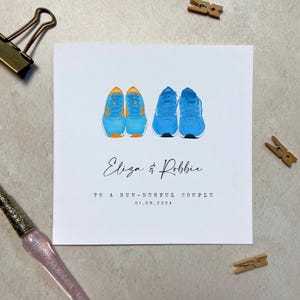 May include: A white card with a blue and orange illustration of two pairs of running shoes. The text "Eliza & Robbie" is written in cursive. The text "To a run-derful couple" and "01.08.2024" is printed below the illustration.