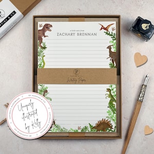 May include: A set of lined writing paper in a brown box, featuring watercolour dinosaur illustrations and green foliage. The paper includes the text 'a little note from ZACHARY BRENNAN'. A brown paper band reads 'POPPINS & CO. Writing Paper'.