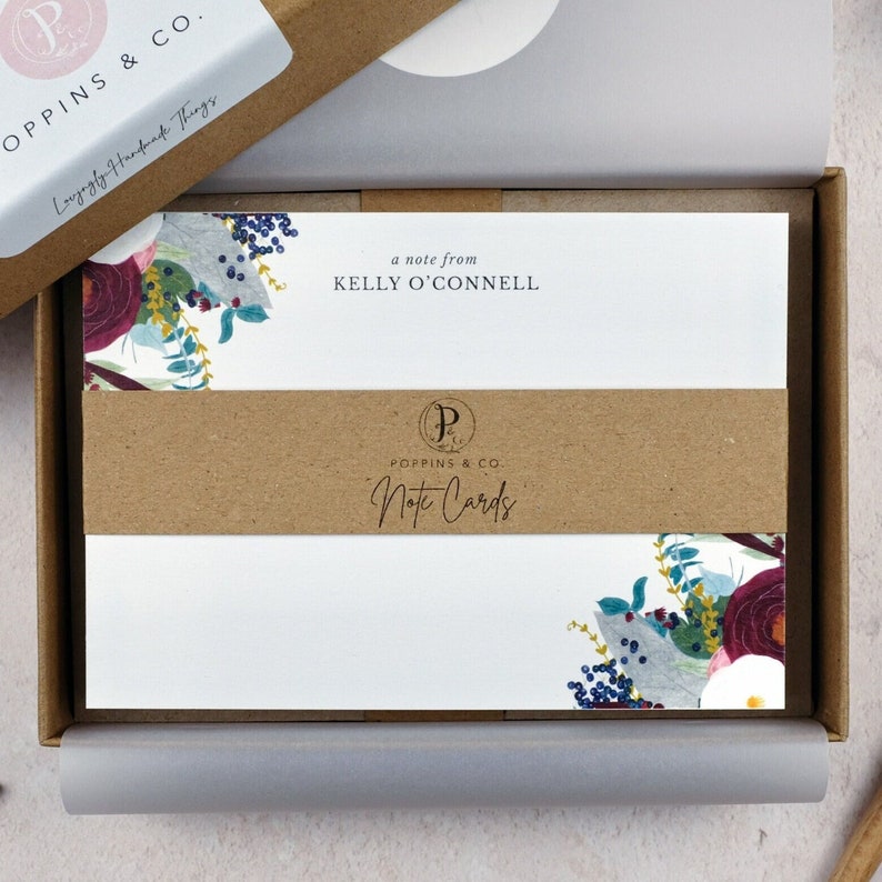 May include: A box of note cards with a floral design in shades of blue, purple, and red. The box is brown and has the text "a note from KELLY O'CONNELL" and "POPPINS & CO. Note Cards" printed on it.