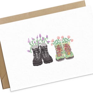 May include: Watercolor illustration of two pairs of hiking boots, one pair in black and the other in green with floral patterns. Purple and pink flowers sprout from the tops of the boots, set against a white background.