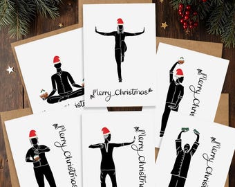 May include: A set of Christmas cards featuring black silhouettes of people in various yoga poses, each wearing a Santa hat. The cards are white with the words "Merry Christmas" in script. The cards are on a wooden surface.