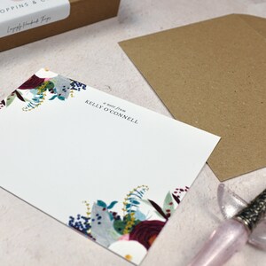 May include: A white note card with a floral border in shades of blue, red, and green. The text "a note from KELLY O'CONNELL" is printed in black at the top of the card.