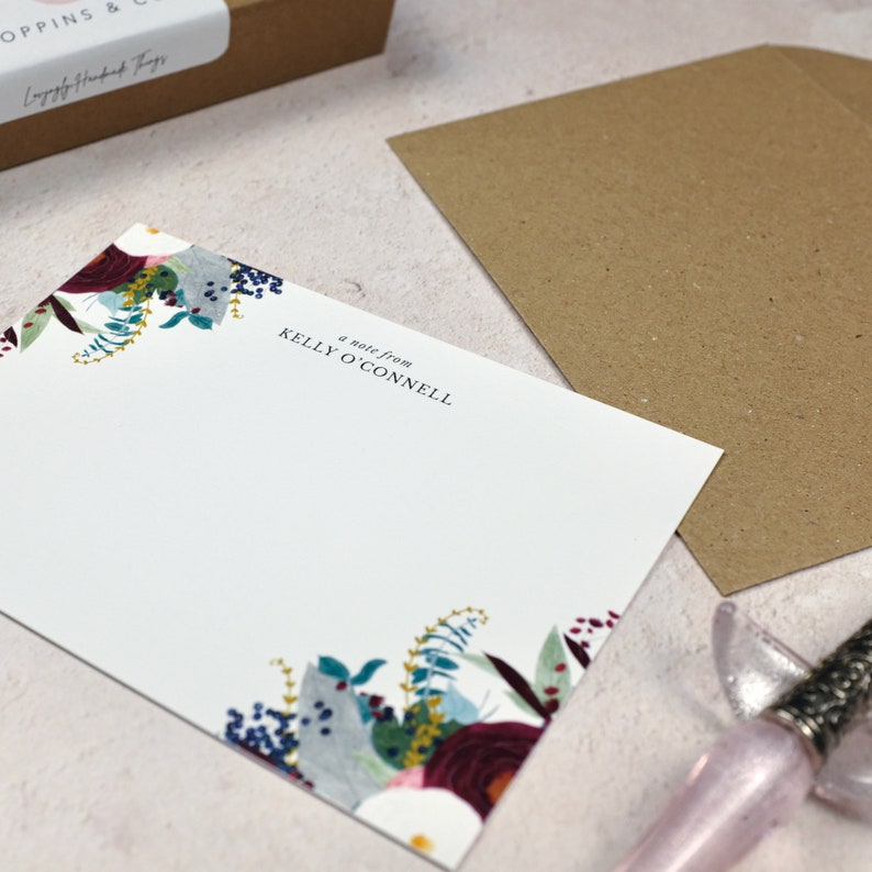 May include: A white note card with a floral border in shades of blue, red, and green. The text "a note from KELLY O'CONNELL" is printed in black at the top of the card.