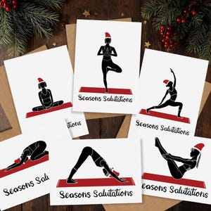 May include: A collection of white greeting cards featuring black silhouettes of yoga poses, each wearing a red Santa hat. The cards are printed with the text "Seasons Salutations" in black. The cards are on a wooden surface with holiday decorations.