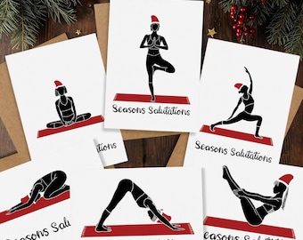 May include: A collection of white greeting cards featuring black silhouettes of yoga poses, each wearing a red Santa hat. The cards are printed with the text "Seasons Salutations" in black. The cards are on a wooden surface with holiday decorations.