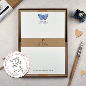 May include: A boxed stationery set featuring white writing paper with a blue butterfly design and the text "a letter from JENNY HIRONS." A brown band reads "Writing Paper." A round sticker says "Uniquely illustrated by Kelly." The set is in a brown box.