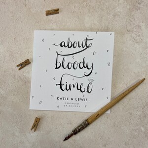 K&ouml;nnte beinhalten: Eine quadratische wei&szlig;e Karte mit schwarzer Kalligraphie, die "about bloody time" liest, mit einer Diamantring-Illustration. Die Karte ist f&uuml;r "KATIE & LEWIS", verlobt am 09.03.2024. Kleine Herz-Doodles umgeben den Text. Ein Holzstift und drei kleine W&auml;scheklammern sind ebenfalls sichtbar.