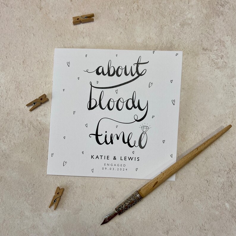 K&ouml;nnte beinhalten: Eine quadratische wei&szlig;e Karte mit schwarzer Kalligraphie, die "about bloody time" liest, mit einer Diamantring-Illustration. Die Karte ist f&uuml;r "KATIE & LEWIS", verlobt am 09.03.2024. Kleine Herz-Doodles umgeben den Text. Ein Holzstift und drei kleine W&auml;scheklammern sind ebenfalls sichtbar.