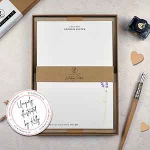 以下が含まれることがあります： 茶色の箱に入った文房具セット。セットには、「a letter from GEORGIA FOSTER」と「Writing Paper」の文字が入った白い便箋が含まれています。下のシートにはラベンダーの小枝が描かれています。ペン、インクボトル、ハート型の切り抜きも含まれています。