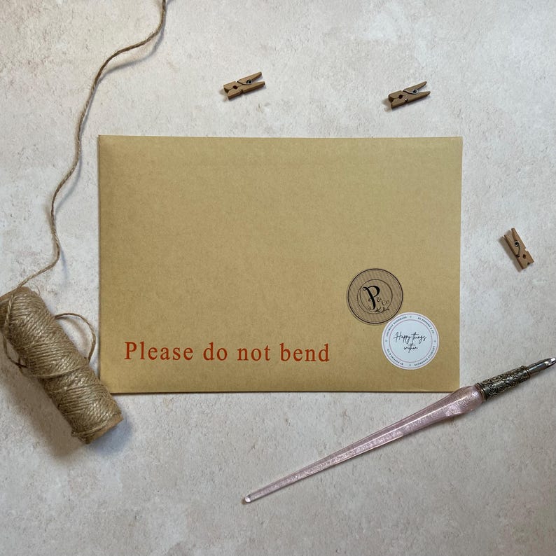 May include: A brown paper envelope with the text "Please do not bend" in red. The envelope has two circular stickers and is accompanied by a pink pen, twine, and small wooden clothespins.