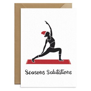 May include: A white greeting card with a brown envelope features a black silhouette of a person in a yoga pose, wearing a Santa hat. The card rests on a red yoga mat, with the words "Seasons Salutations" in black script.