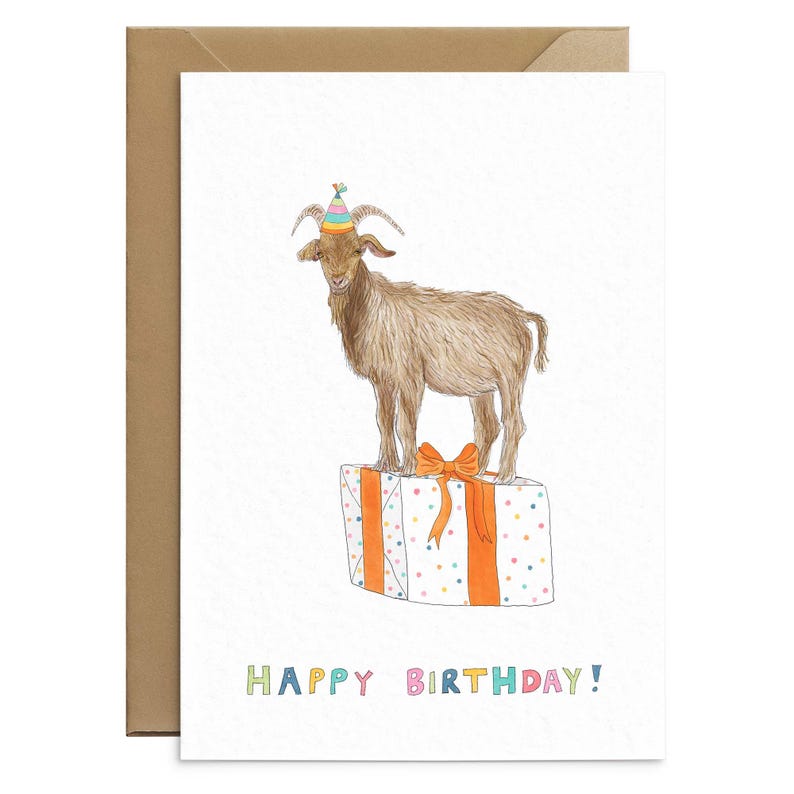 May include: A birthday card featuring a brown goat wearing a colorful party hat standing on top of a white gift box with orange ribbon and colorful confetti. The text "HAPPY BIRTHDAY!" is written in colorful letters at the bottom of the card.