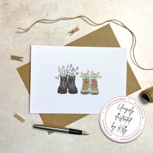 May include: A greeting card featuring watercolor illustrations of hiking boots filled with flowers. One pair is black, the other green. The card is on a white background, with a brown envelope, pen, and a sticker that says "Uniquely illustrated by Kelly."