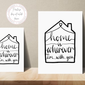 May include: Two white art prints with black text that reads "Home is wherever I'm with you" inside a house outline. The prints are on a wooden surface. A circular logo with text is in the upper left corner.