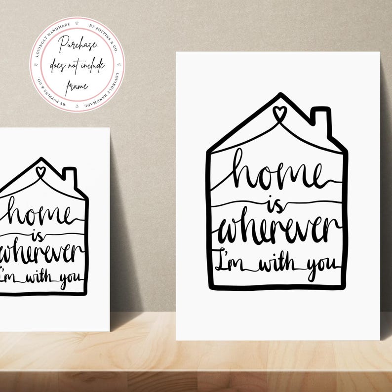 May include: Two white art prints with black text that reads "Home is wherever I'm with you" inside a house outline. The prints are on a wooden surface. A circular logo with text is in the upper left corner.
