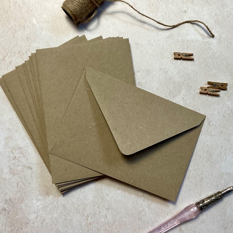 May include: A stack of kraft paper envelopes with matching cards, a ball of twine, and small wooden clothespins. A pink pen with a decorative silver-colored metal tip is also visible. The envelopes are triangular and the cards are rectangular.