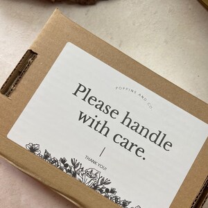 May include: A white sticker on a brown cardboard box with the text "Please handle with care." and the company name "Poppins and Co." below. The sticker has a black floral design at the bottom.