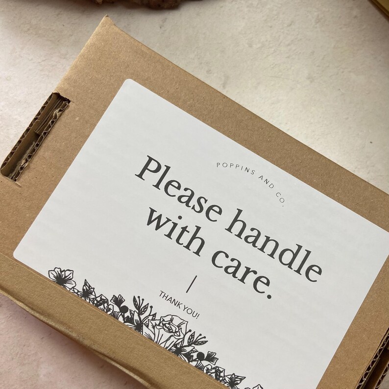 May include: A white sticker on a brown cardboard box with the text "Please handle with care." and the company name "Poppins and Co." below. The sticker has a black floral design at the bottom.