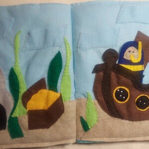 May include: A felt book open to a colorful underwater scene. The left page shows a cave, seaweed, and treasure. The right page features a sunken ship with a diver. The background is light blue.