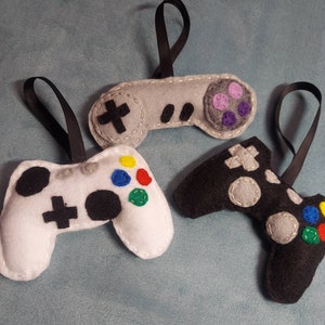May include: Three felt video game controller ornaments. One is gray with purple buttons, one is white with black buttons and colorful accents, and one is black with gray buttons and colorful accents.
