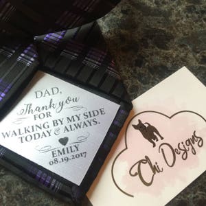 Peut inclure: Une carte blanche avec du texte noir qui dit "DAD, Thank you FOR WALKING BY MY SIDE TODAY & ALWAYS. EMILY 08.19.2017" est attachée à une cravate à motif noir et violet. Une carte rose avec un cœur doré et le texte "Chi Designs" est en arrière-plan.