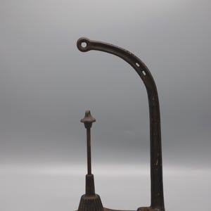 May include: An antique, cast iron bottle capper with a curved arm and a vertical post. The capper is a dark, rusty brown color and sits on a small base. The curved arm has a hole at the end.