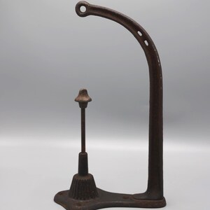 May include: An antique, cast iron bottle capper with a curved arm and a small, decorative post. The capper is a dark, rusty brown color and sits on a rectangular base. The curved arm has two small holes.