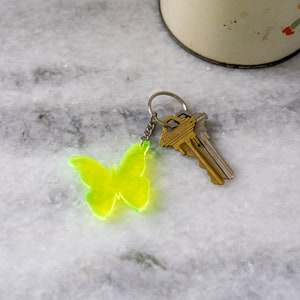 Puede incluir: Un llavero de mariposa verde con dos llaves plateadas unidas. El llavero está sobre una superficie de mármol blanco.