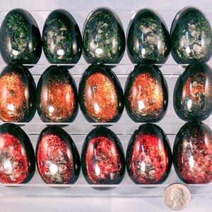 May include: A set of 15 black eggs with glitter embedded inside. The eggs are arranged in three rows of five. The glitter is in shades of green, orange, and red.