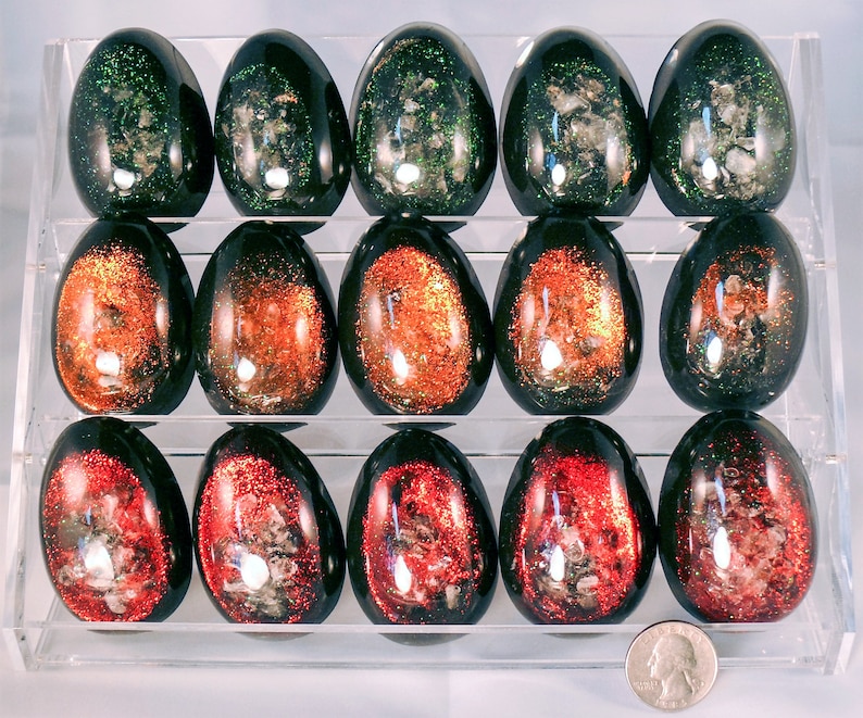 May include: A set of 15 black eggs with glitter embedded inside. The eggs are arranged in three rows of five. The glitter is in shades of green, orange, and red.